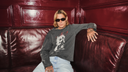 A person wearing a dark gray graphic long-sleeve shirt and light blue jeans sits on a glossy dark red leather couch against a matching paneled wall.